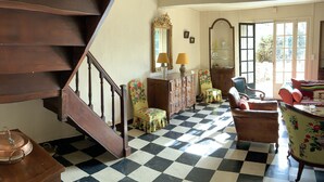 Ein Zimmer mit Schachbrettboden, einer Holztreppe, einem floralen Sessel, einem Holzschrank und einem Spiegel mit goldener Rahmung.
