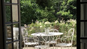 Eine Terrasse im Freien mit einem runden Tisch und weißen Metallstühlen, umgeben von Grünpflanzen.
