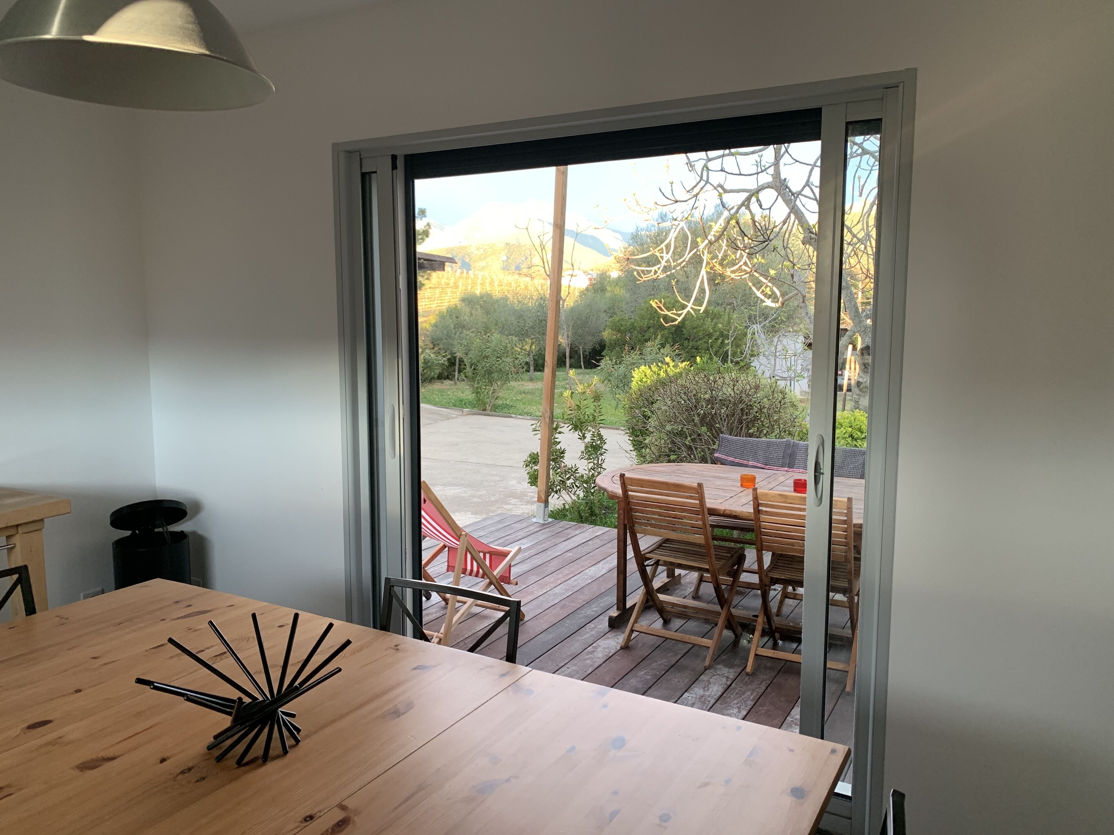 Un tavolo da pranzo in legno con sedie, un piccolo cestino e una lampada a sospensione in una stanza con una porta scorrevole in vetro che dà su un patio esterno con tavolo e sedie.