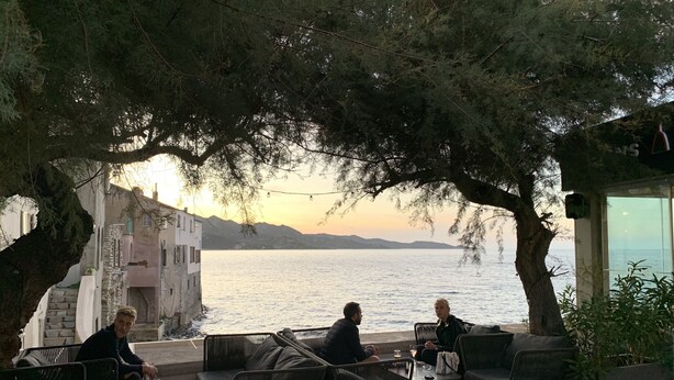 Eine Terrasse mit Sitzgelegenheiten im Freien mit Blick auf ein Gewässer, im Hintergrund Gebäude.