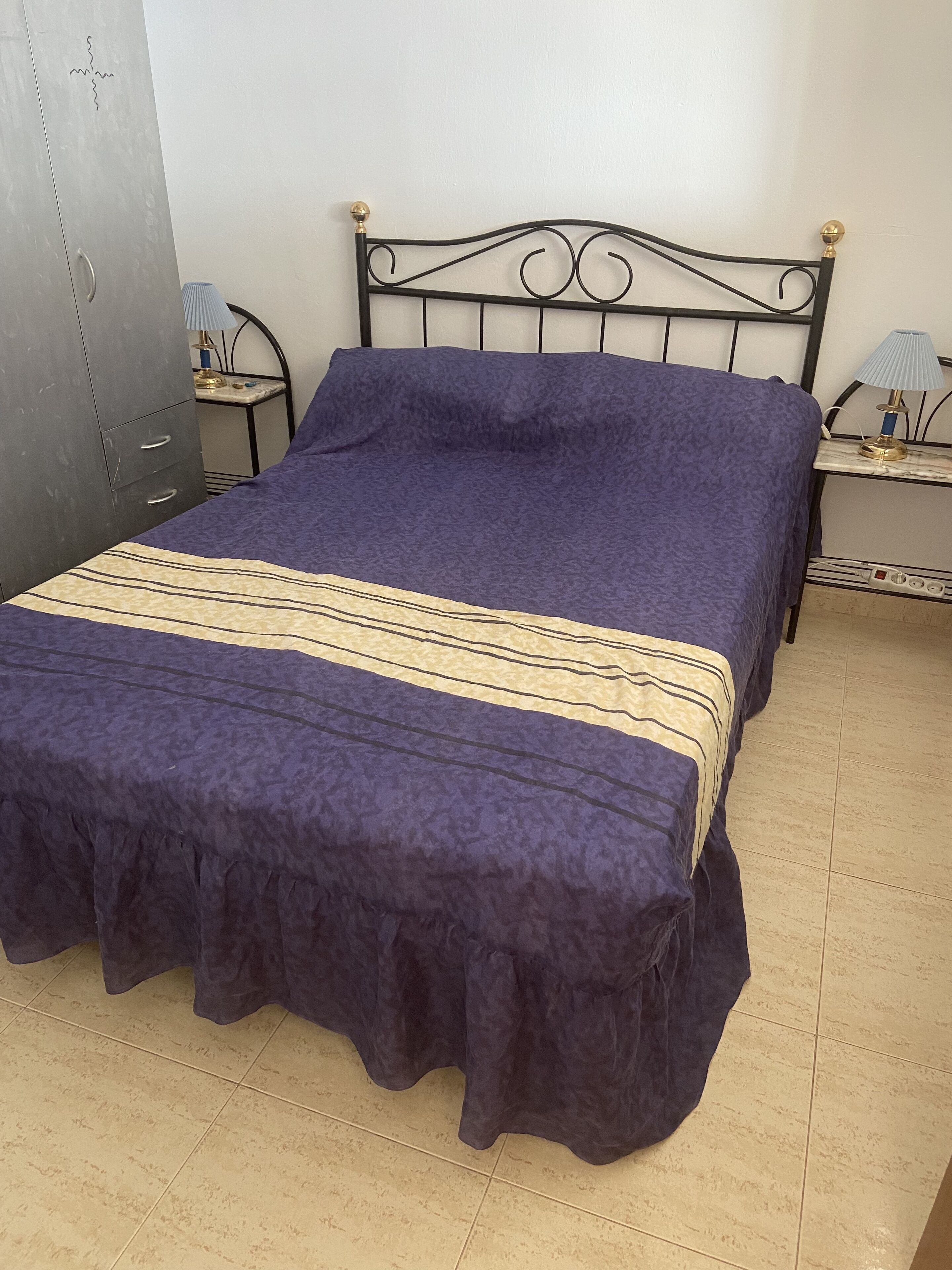 Una cama con una colcha azul oscuro y falda fruncida, cabecero de metal con detalles decorativos y dos mesitas de noche con lámparas.