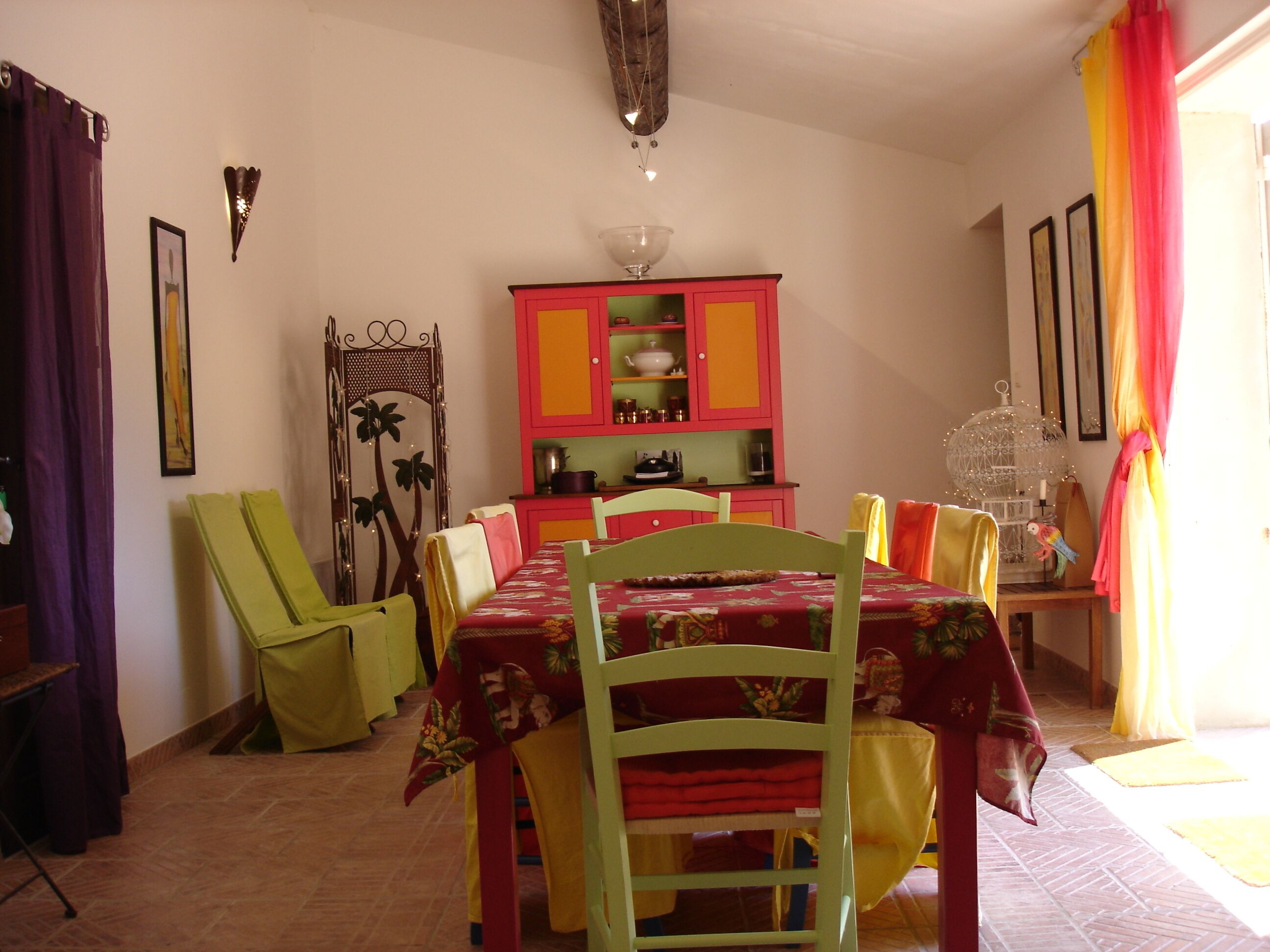 A dining area with a table set for four, surrounded by chairs. A colorful cabinet with dishes is visible in the background.