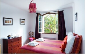 Ein Schlafzimmer mit einem Bett, einer Kommode, einem Fenster mit Vorhängen und gerahmten Bildern an der Wand.