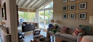 A spacious living area with a sofa, coffee table, and a dining table with chairs. There are framed pictures on the wall and a view of the outdoors through large windows.