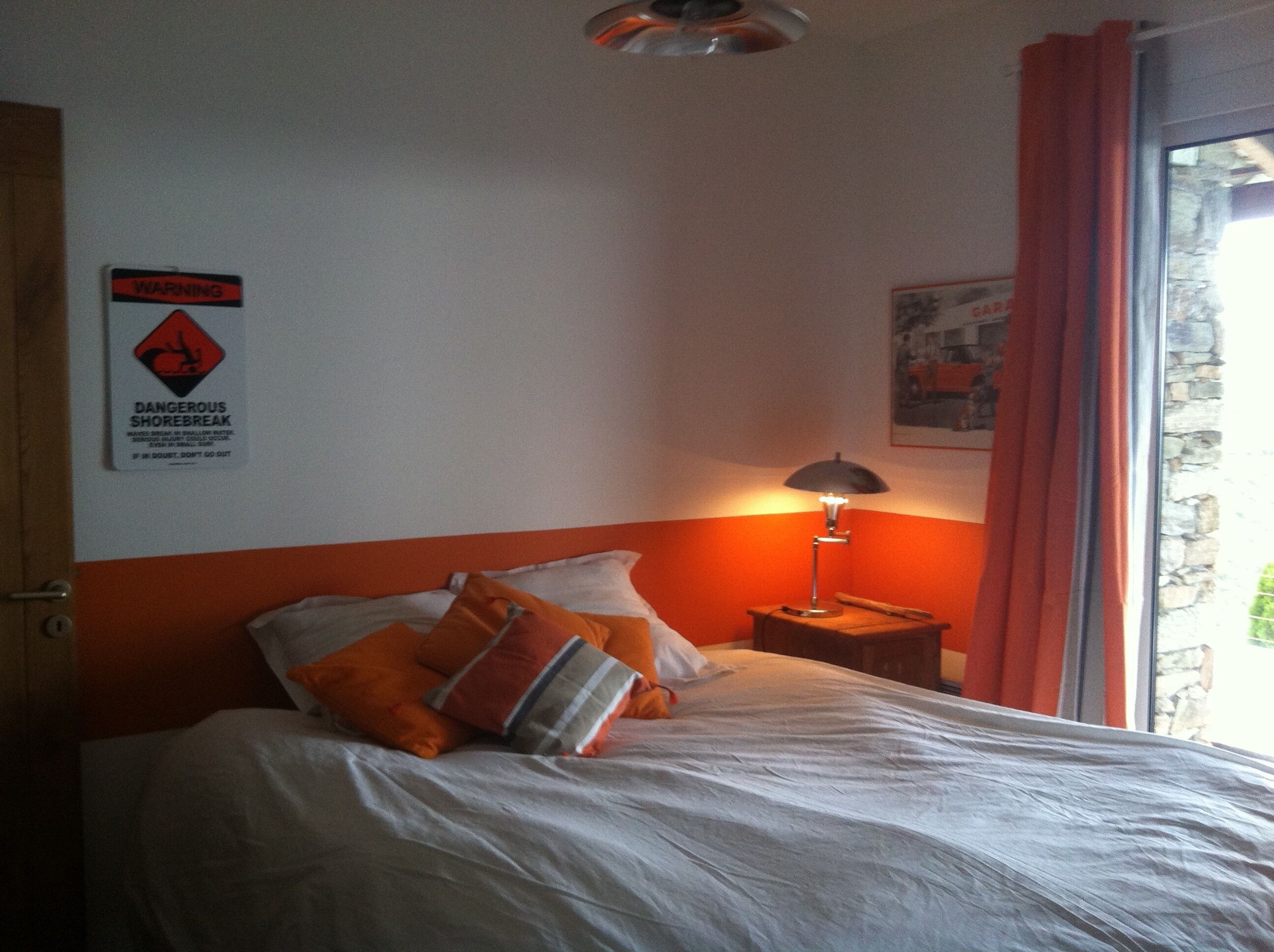 Ein Schlafzimmer mit einem Bett, einem Nachttisch mit Lampe, einem Warnschild und einem Fenster mit Vorhängen.