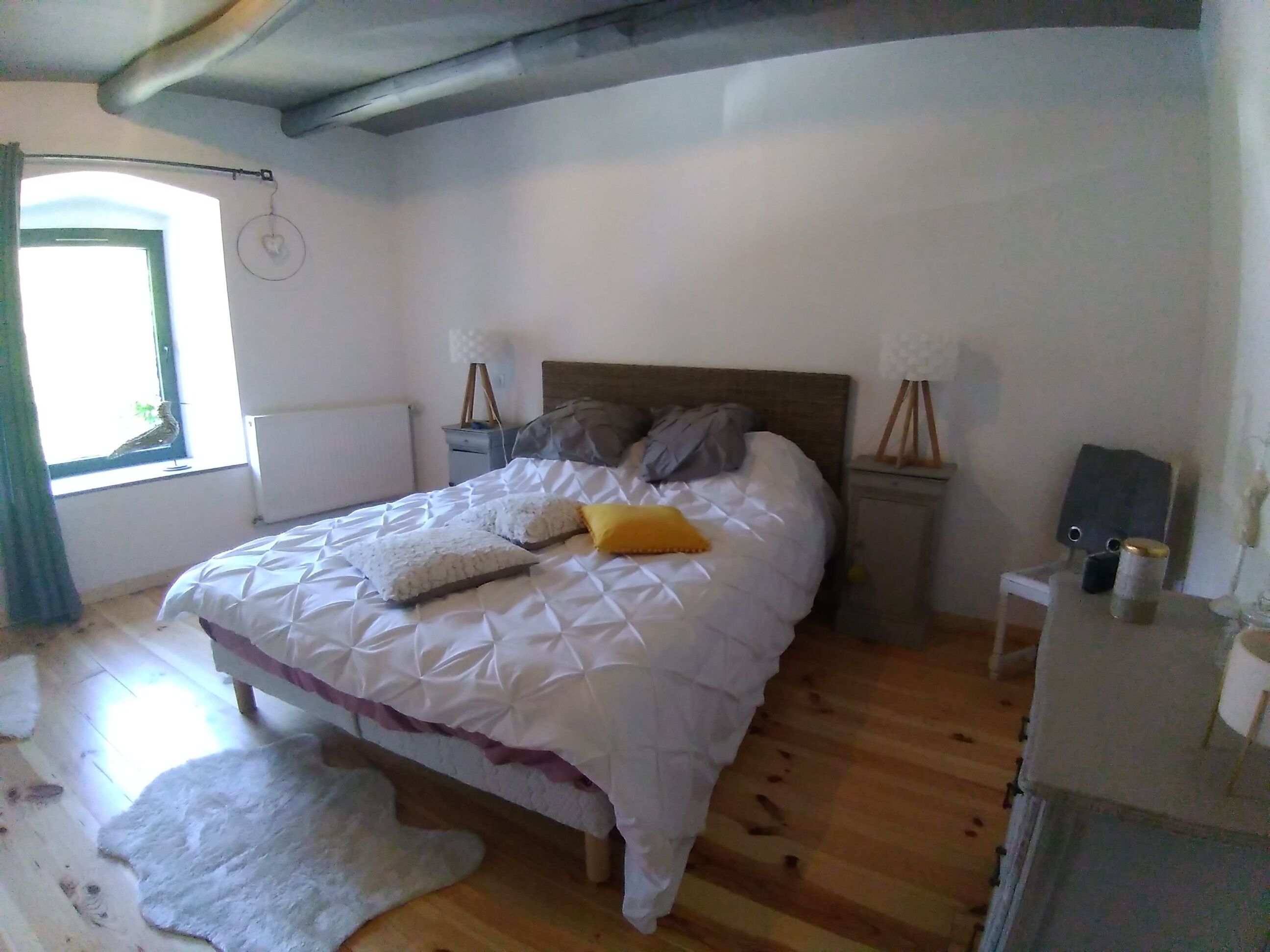 A bedroom with a bed, wooden floor, a white rug, a window with curtains, and a radiator.