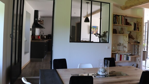 Une cuisine moderne avec une hotte noire, un coin repas avec une table et des chaises en bois, et une bibliothèque remplie de livres et d’objets décoratifs.