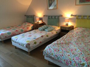 Quarto com três camas individuais, cada uma com colcha e almofada estampadas com padrão floral. Há mesas de cabeceira com luminárias, e o quarto possui teto inclinado.