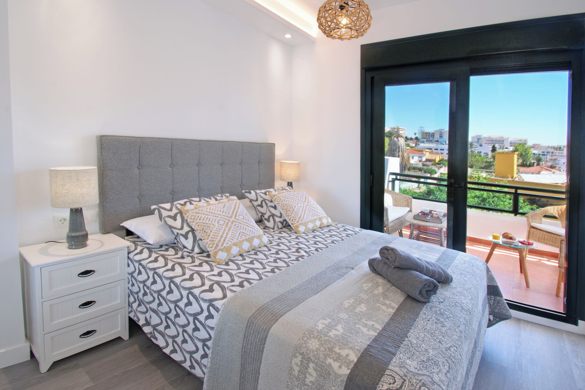 Une chambre moderne avec une tête de lit capitonnée grise, une literie à motifs, une table de chevet avec une lampe et un balcon offrant une vue sur la ville.