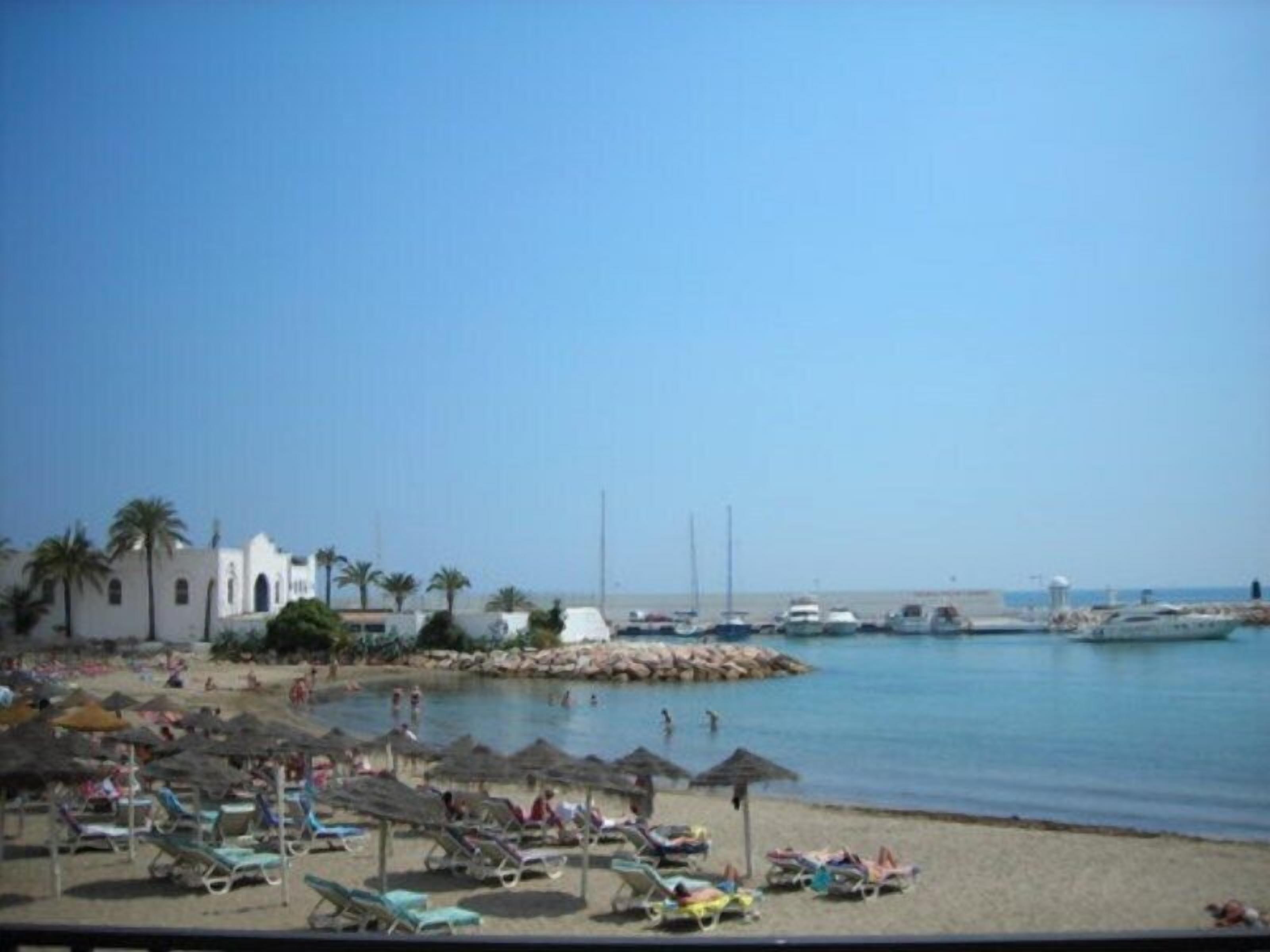 A beach with sun loungers and umbrellas, a rocky breakwater, and boats in the water.