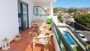 Ein Balkon mit Korbmöbeln, einem Glastisch mit Frühstücksgebäck, einer kleinen weißen Laterne und Blick auf einen Swimmingpool und eine Straße.