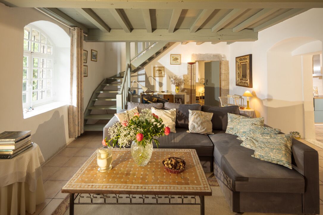 A cozy living room with a sofa, a coffee table with a vase and a muffin, a staircase, and a kitchen area in the background.