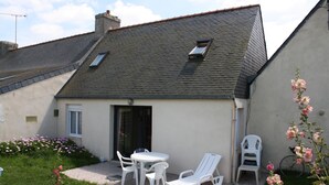 A small house with a sloped roof, a patio with a table and chairs, and a garden with pink flowers. - House                         (Tréguennec)