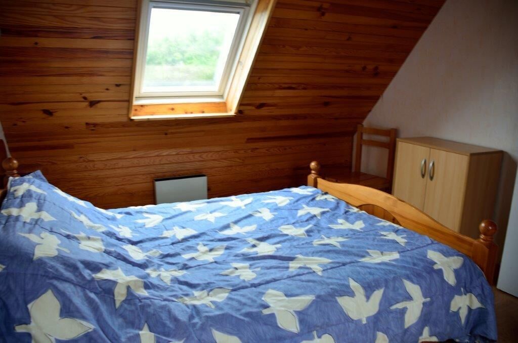 Une chambre avec un plafond en pente, un cadre de lit en bois, une couvre-lit bleue à motifs blancs, une petite fenêtre et une armoire en bois.