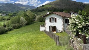 Una casa de piedra blanca con tejado rojo, rodeada de campos verdes exuberantes y montañas al fondo.