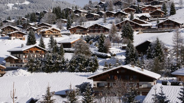 Een besneeuwde bergdorp met houten chalets, elk met balkons en grote ramen, omringd door bomen en een heldere lucht.
