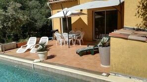 Uma área à beira da piscina com mesa, cadeiras, plantas em vasos e um guarda-sol branco.