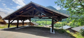 A wooden pavilion with a sloped roof, surrounded by a rural landscape with houses and mountains in the background.