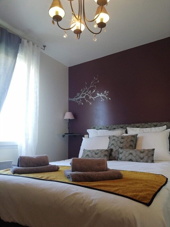 A hotel room with a large bed, a chandelier, a wall art of a tree, and a window with curtains. - The Door Of The Great Blue (binic)