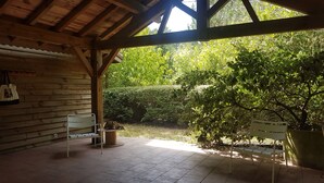 Eine überdachte Terrasse mit Holzboden, einem weißen Stuhl, einem Tisch mit einer Topfpflanze und Blick auf einen Garten mit Hecke und Bäumen.
