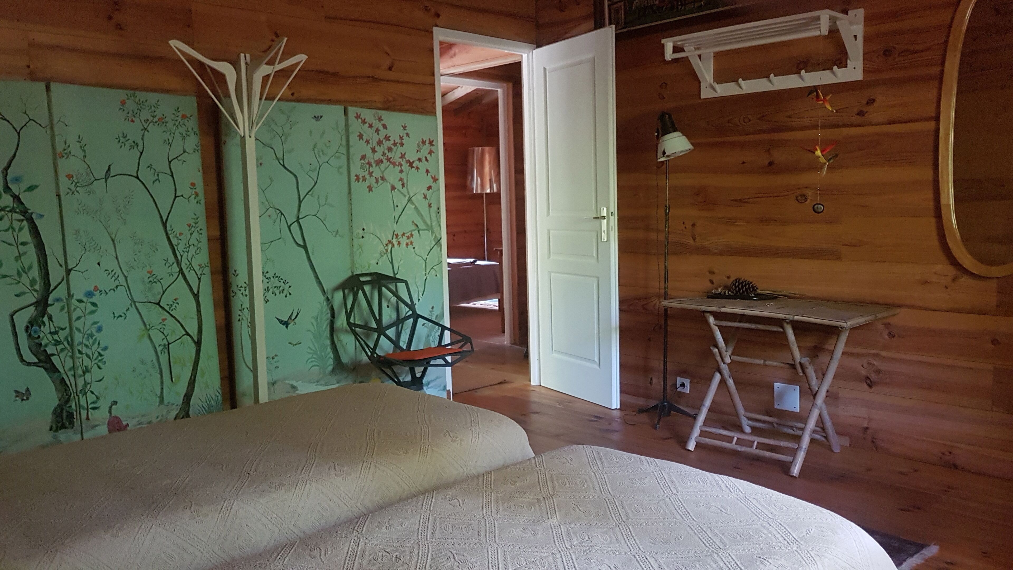 Een houten kamer met een bed, een stoel, een tafel en een deur die naar een andere kamer leidt.