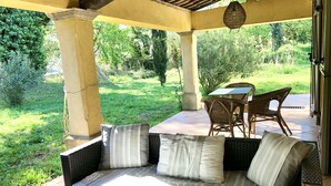 A covered outdoor seating area with a wicker sofa and chairs, a table, and a view of a lush garden. - Single storey villa, 4 bedrooms, swimming pool, near the beaches of the Gulf of Saint Trop (Grimaud)