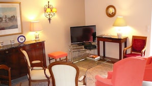 Una sala de estar con televisor, sillas de madera, un sillón rojo y una mesita con lámpara.