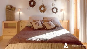 Quarto com cabeceira de madeira, cama com colcha marrom, várias almofadas, duas mesas de cabeceira com abajures e um cesto do lado esquerdo.