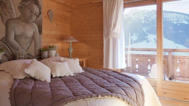Quarto de cabana de madeira com cama, almofadas, luminária e vista para uma paisagem coberta de neve.