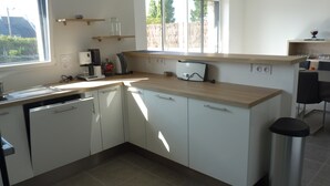 A modern kitchen with white cabinets, a dishwasher, a coffee maker, and a toaster. There are wooden shelves above the counter and a window providing natural light. - Carnac - Close to beaches and shops - 3 bedrooms (Carnac)