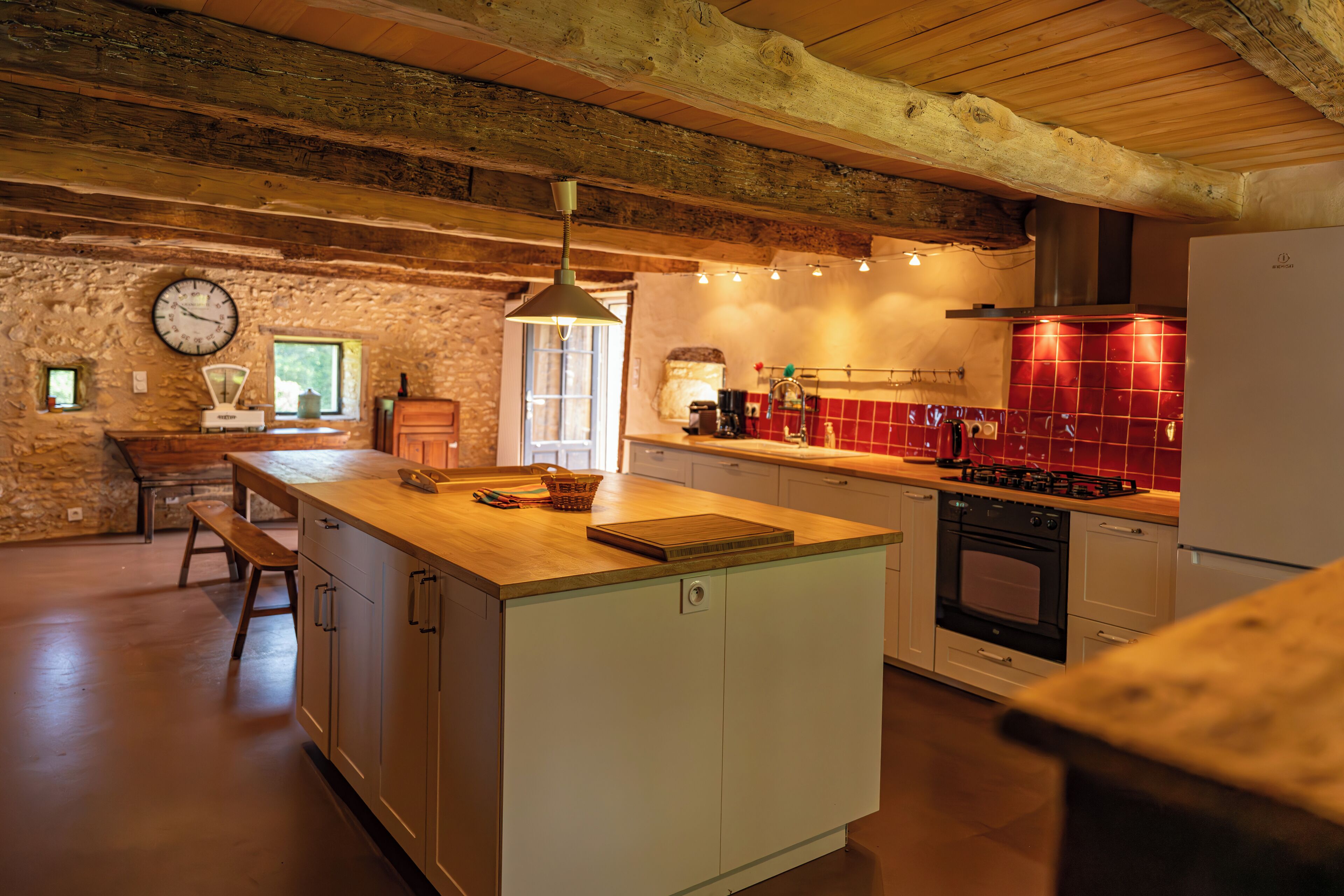 A rustic kitchen with wooden beams, a stone wall, a wooden bench, and a red-tiled backsplash.