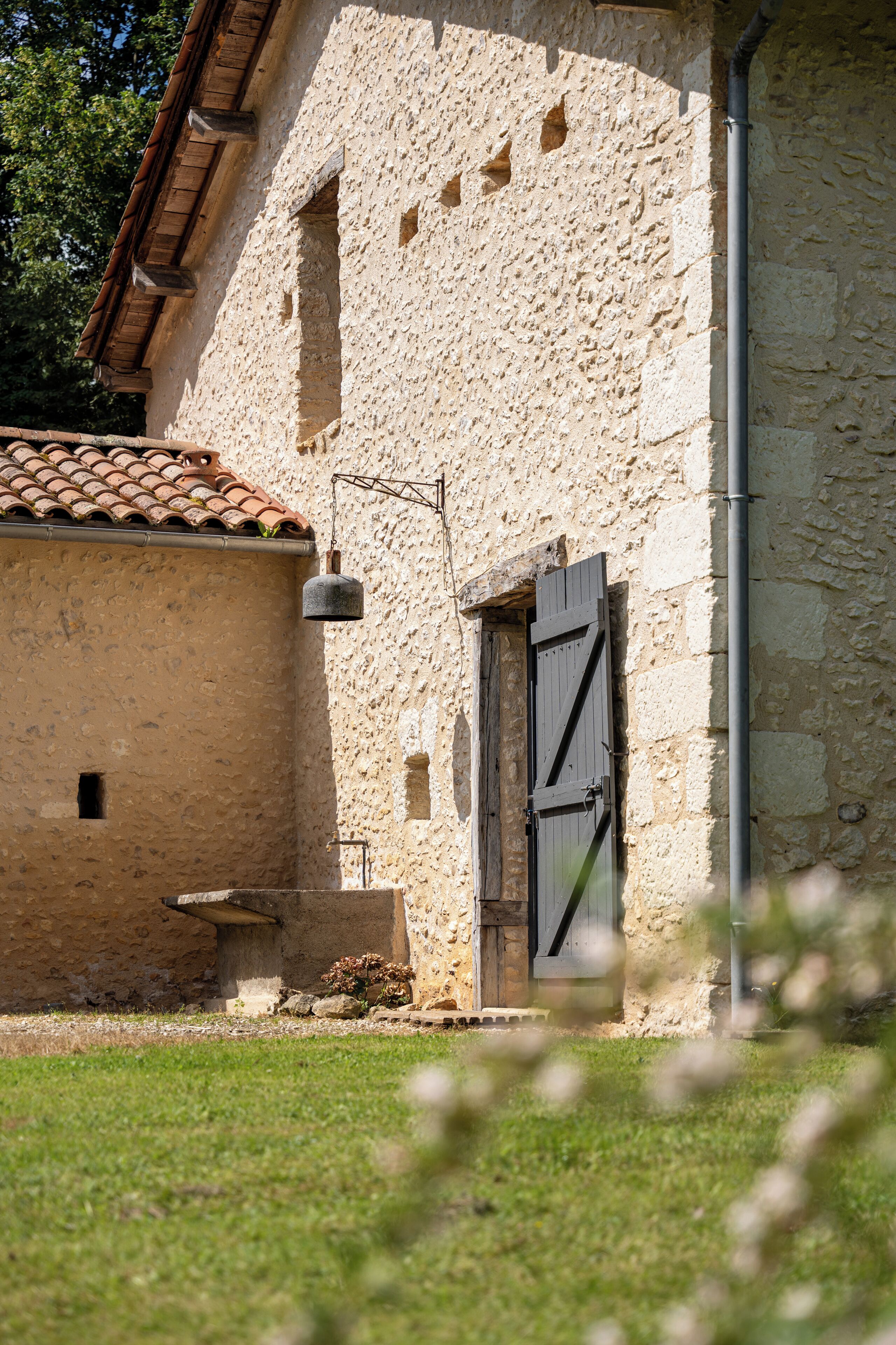 A rustic stone building with a tiled roof, a bell, and a wooden door.