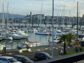 A marina with numerous boats docked, a cityscape in the background, and a street with cars and a scooter. - GREAT 2-BEDROOM APARTMENT WITH AIR CONDITIONING, HARBOR VIEW, PARKING, BEACH 50 m AWAY, SWIMMING POOL, WIFI, SPAIN (ferry service). (Hendaye)