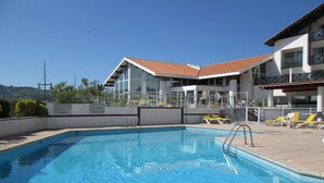 Une piscine à l’eau d’un bleu limpide, entourée de chaises longues et bordée d’un bâtiment dont le toit est recouvert de tuiles.