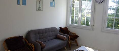 A living room with a grey sofa, two wicker chairs, a small wooden table with a bowl of fruit, and a window with a clock.