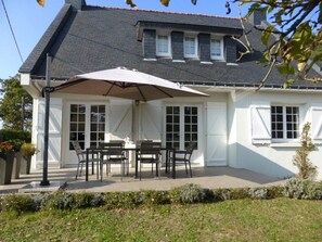 Une maison dotée d’un patio couvert comprenant une table et des chaises, un grand parasol, et un jardin en premier plan.