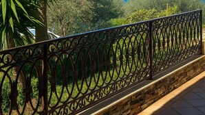 Ein Balkon mit schmiedeeisernem Geländer, der auf eine Küstenlandschaft mit Bäumen und dem Meer blickt.