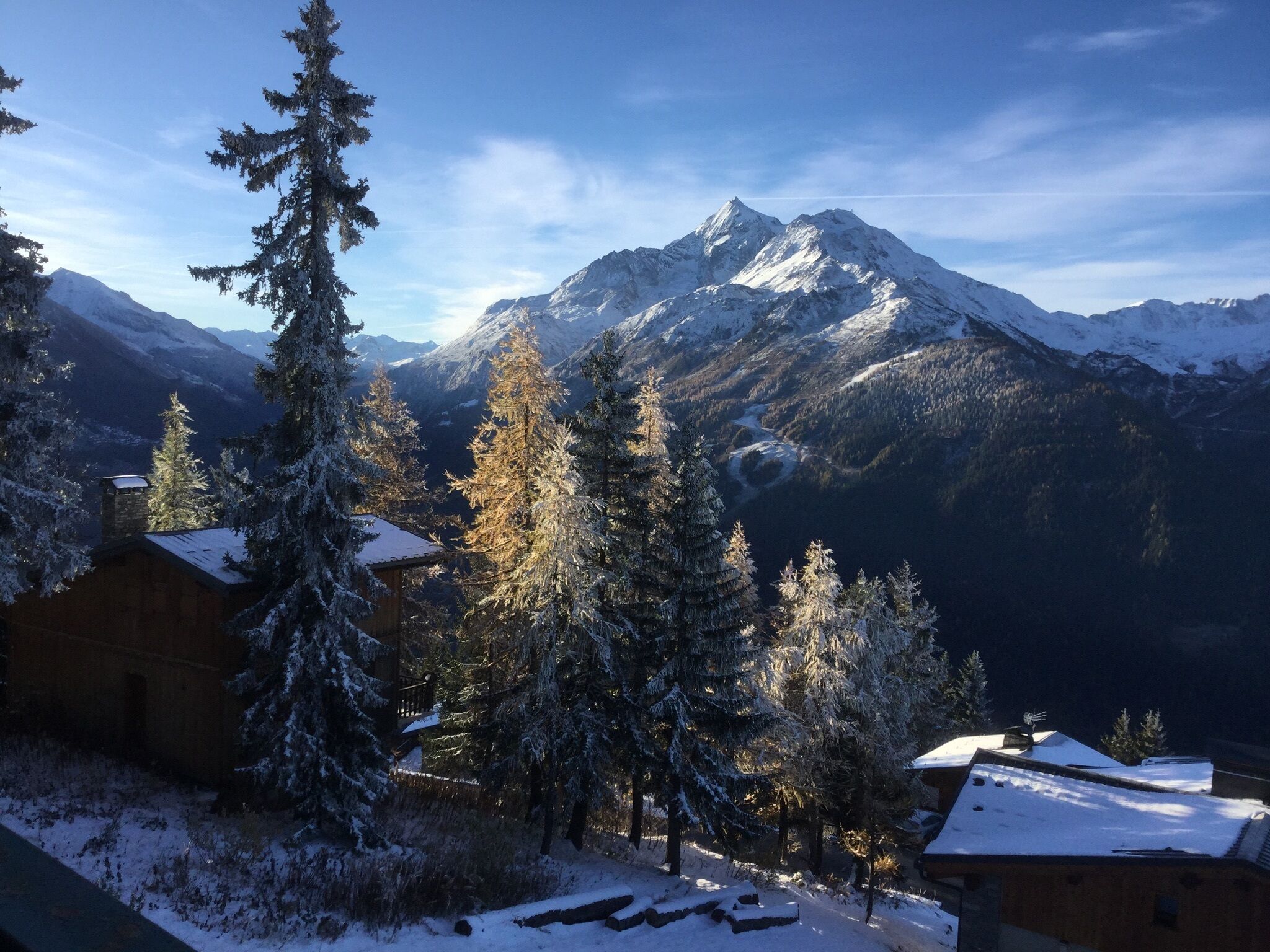 A mountain cabin with a snow-covered roof, surrounded by evergreen trees and a view of a snow-capped mountain range.