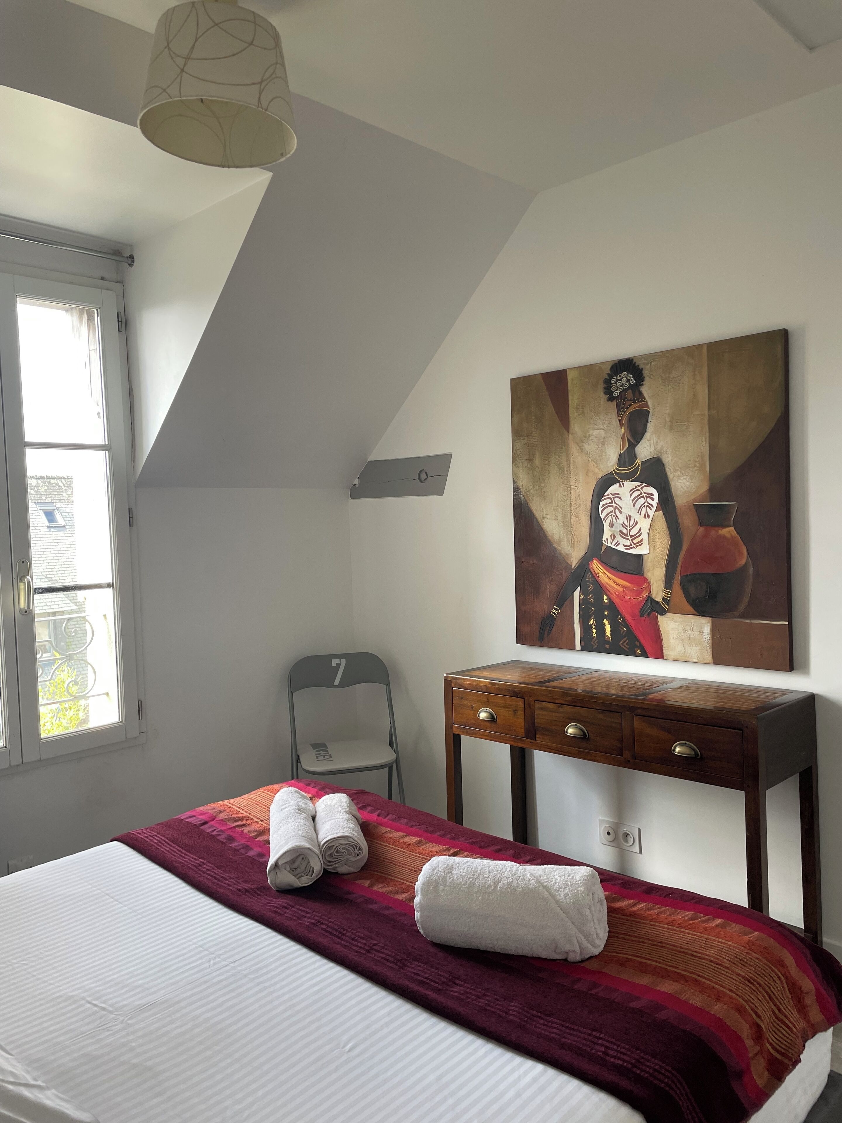 A bedroom with a bed, a chair, a desk, a painting, and a window.