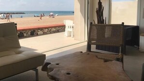 Une pièce dotée d’une grande fenêtre donnant sur une plage, d’une table en bois, d’un canapé blanc et d’un fauteuil en osier.