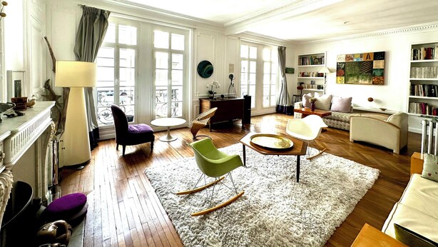 A spacious living room with a fireplace, a large window with curtains, a bookshelf, and a dining area with a round table.