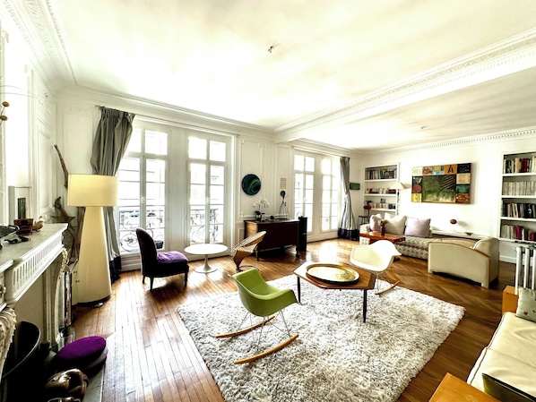 A spacious living room with a fireplace, a large window with curtains, a bookshelf, and a dining area with a round table.