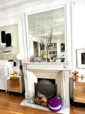 A room with a fireplace, a large mirror, a radiator, a wooden floor, and a bookshelf.
