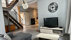 A modern living room with a staircase, a large wall clock, a flat-screen TV, and a sofa with patterned cushions.