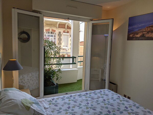 A hotel room with a bed, a nightstand, a lamp, a sliding glass door leading to a balcony with a plant and a view of the building's exterior.