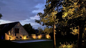 A stone building with a sloped roof, a swimming pool, and a well-lit garden area at dusk. - Luxury property with pool in Tourraine (Saché)