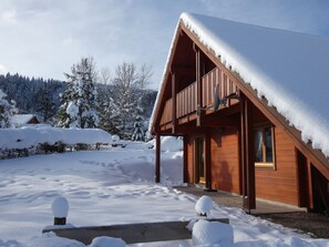 A wooden cabin with a sloped roof covered in snow, surrounded by a snowy landscape and trees.