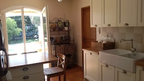 A kitchen with a wooden table, a chair, a window with a view of greenery, and a sink with a vintage faucet. - Beautiful Villa (Bastide) close to the sea and Pic Saint Loup (Sussargues)