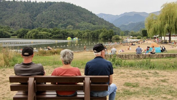 Un lago con gente sentada en bancos y un área verde con árboles y montañas al fondo.