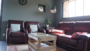 Ein Wohnzimmer mit braunem Ledersofa, zwei Ledersesseln, einem Couchtisch mit Glasplatte, einer Ziegelwand, einem Fenster und einer Uhr.